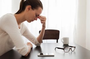 woman pressing finders to bridge of nose looking stressed with phone and glasses on table